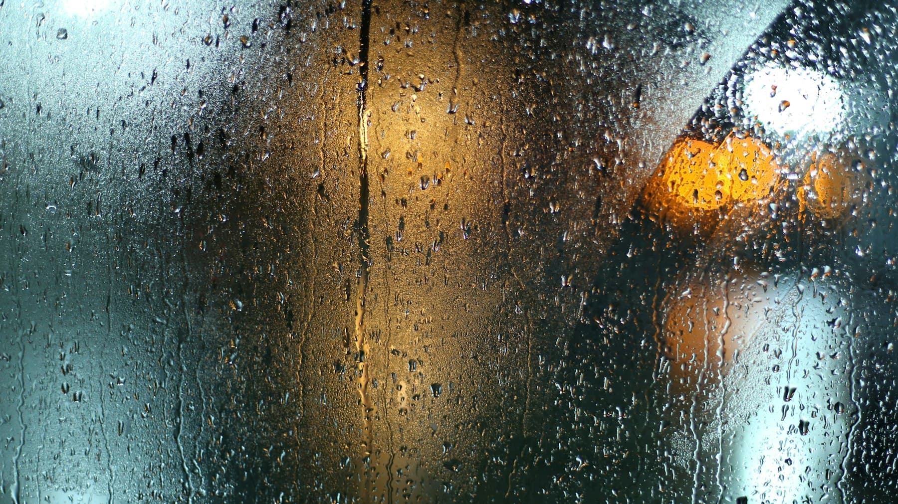 Rain streaking down a window at night with warm street lights blurred behind the glass